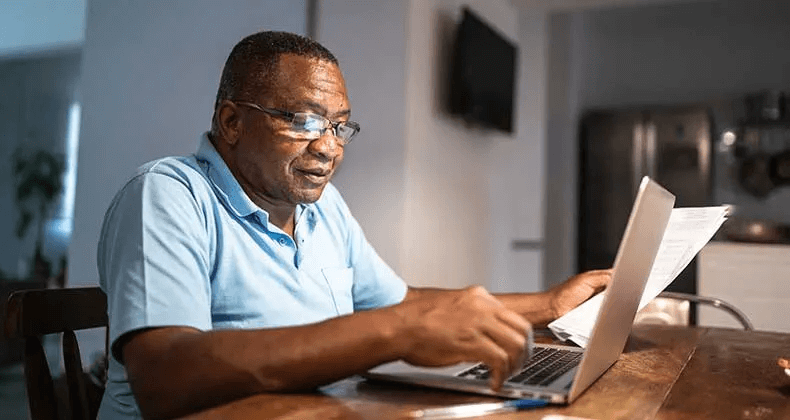 A senior sits at a desk with a laptop and papers