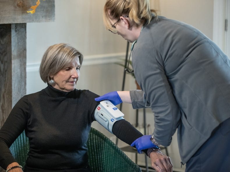 an older woman sits in a chair at home while a nurse checks her blood pressure