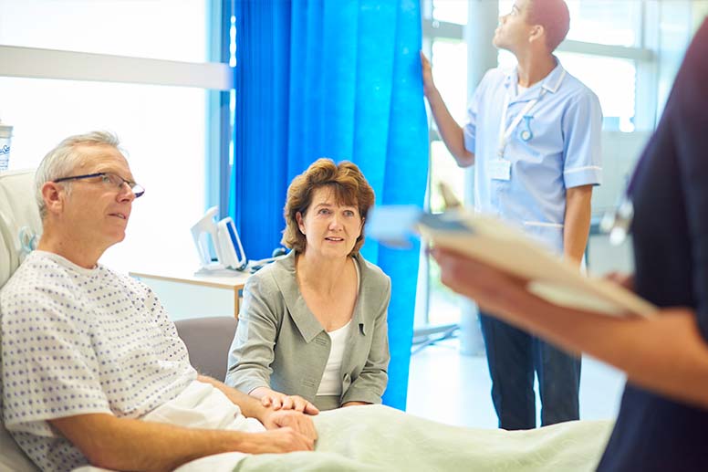 husband in hospital bed holding wife's hand talking to a doctor