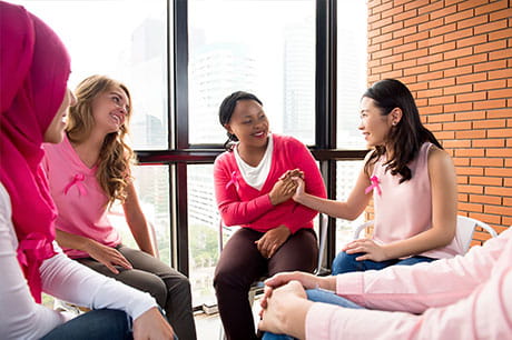 5 women in pink sitting in a circle with breast cancer ribbons pinned to their shirts