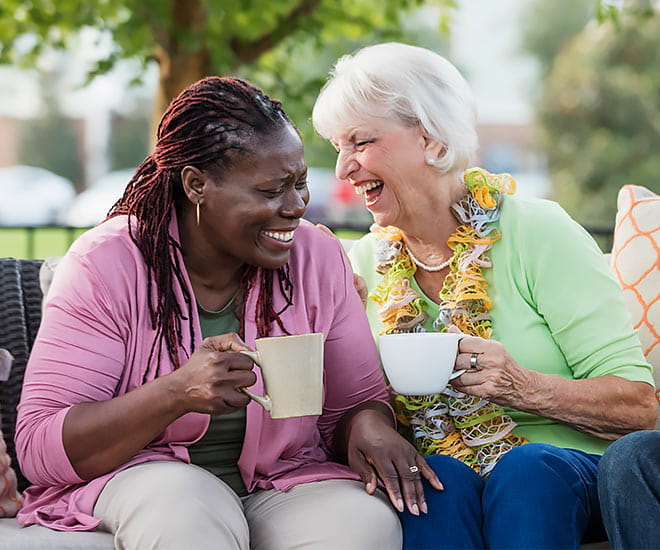 Two women on a park bench having a laugh while drinking coffee or tea.