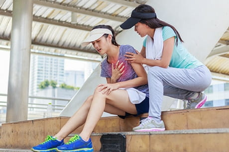 Two women in workout outfits sit on the steps. One is clutching her heart. The other tries to help.