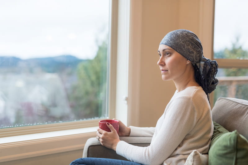 Beautiful breast cancer survivor sits by window wearing headscarf drinking tea.