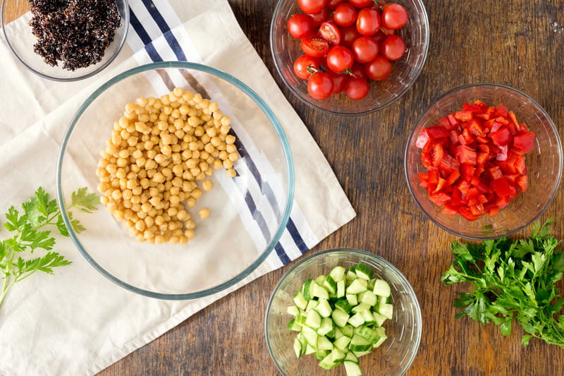 Fresh ingredients in glass bowls on a table including chickpeas, tomato and cucumber.