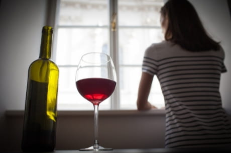 Women looks out of a window with a nearly empty wine bottle and glass of red wine on the table behind her.