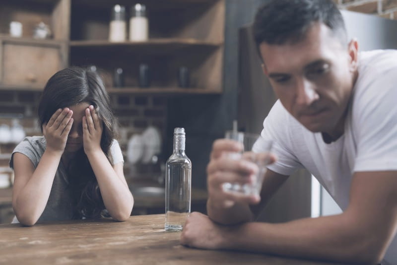 A little girl sits at a table and covers her face with her hands and cries while her father sits and drinks, turning away from her with shame.