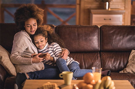 Happy African American mother and daughter relaxing on sofa in the living room and using mobile phone to access myGeisinger.