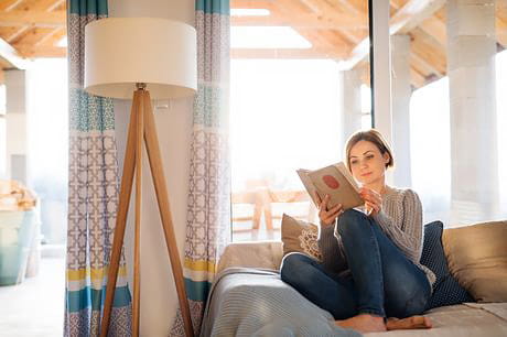 Young woman sitting indoors on a couch at home, reading a book.