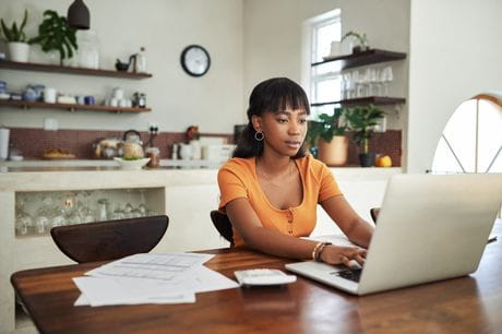 A young woman working from a laptop in her kitchen.