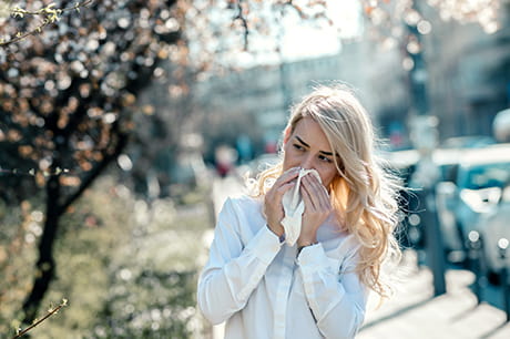 Woman with a runny nose due to allergies, walking outside and blowing nose.