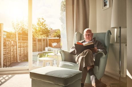 Older man sitting on a couch near a window reading a book. 