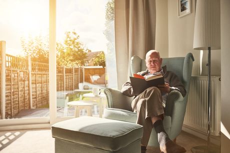 Older man sitting on a couch near a window reading a book.