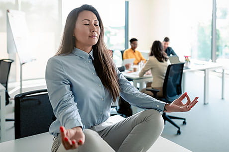 A woman meditates on a table in an office setting while a meeting is being held.