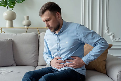 Man in gastric distress is seated on couch while holding his stomach.