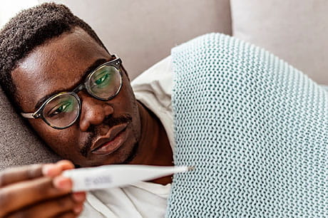 An adult male with flu symptoms checks his temperature as he rests on a couch.
