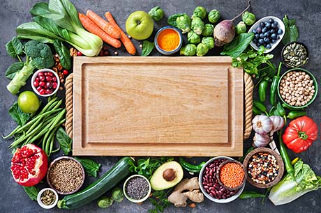 A bounty of fruits, nuts, and vegetables surrounds a large cutting board.