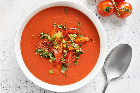 A delicious bowl of gazpacho soup is pictured with a spoon and tomatoes.