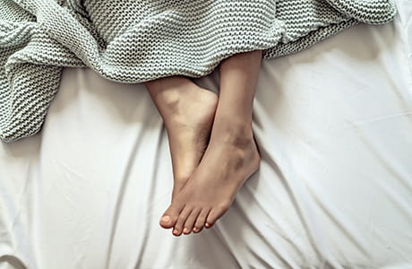 A young woman's legs and feet partially covered by a blanket in bed.