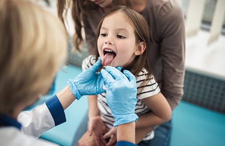 Young girl sitting in her mother's lap, being examined for signs of strep throat by a female healthcare provider.