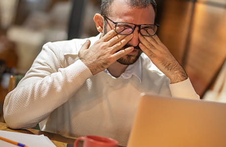 A man rubs his eyes while taking a break from working at a computer.