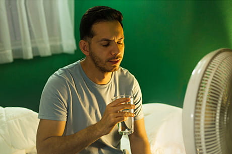 A man tries to cool down from night sweats with a fan and a glass of water.