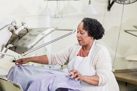 A senior African-American woman in her 60s working at the dry cleaners pressing a shirt with a steam presser. She is an employee, or perhaps the owner of this small business.