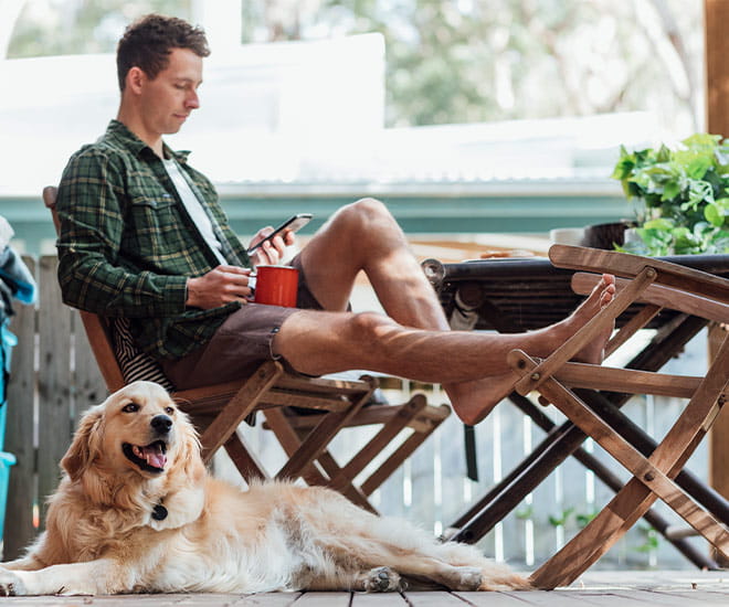 A side view shot of a young man sitting down on a chair outside his house using his smart phone with his dog.