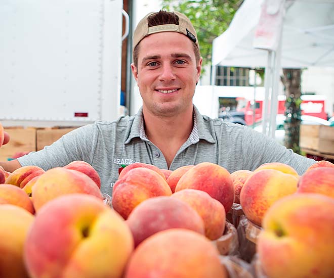 A farmer at a local market stands behind bushels of peaches for sale.