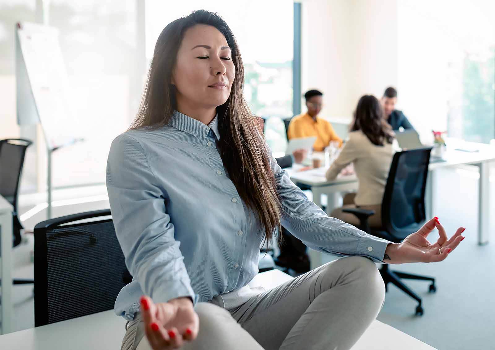 A woman meditates on a table in an office setting while a meeting is being held.