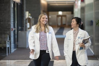 Two female abigail scholars walk in a hallway wearing medical coats