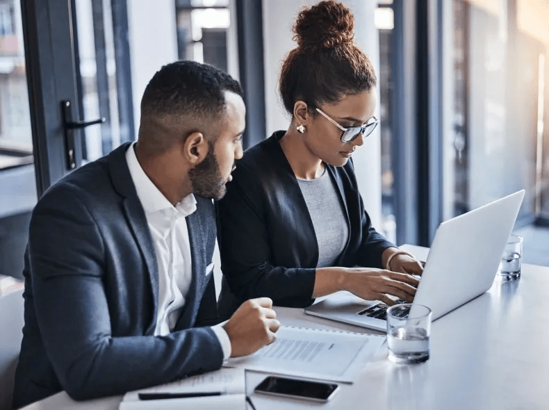 Two office workers in a meeting room consult a laptop