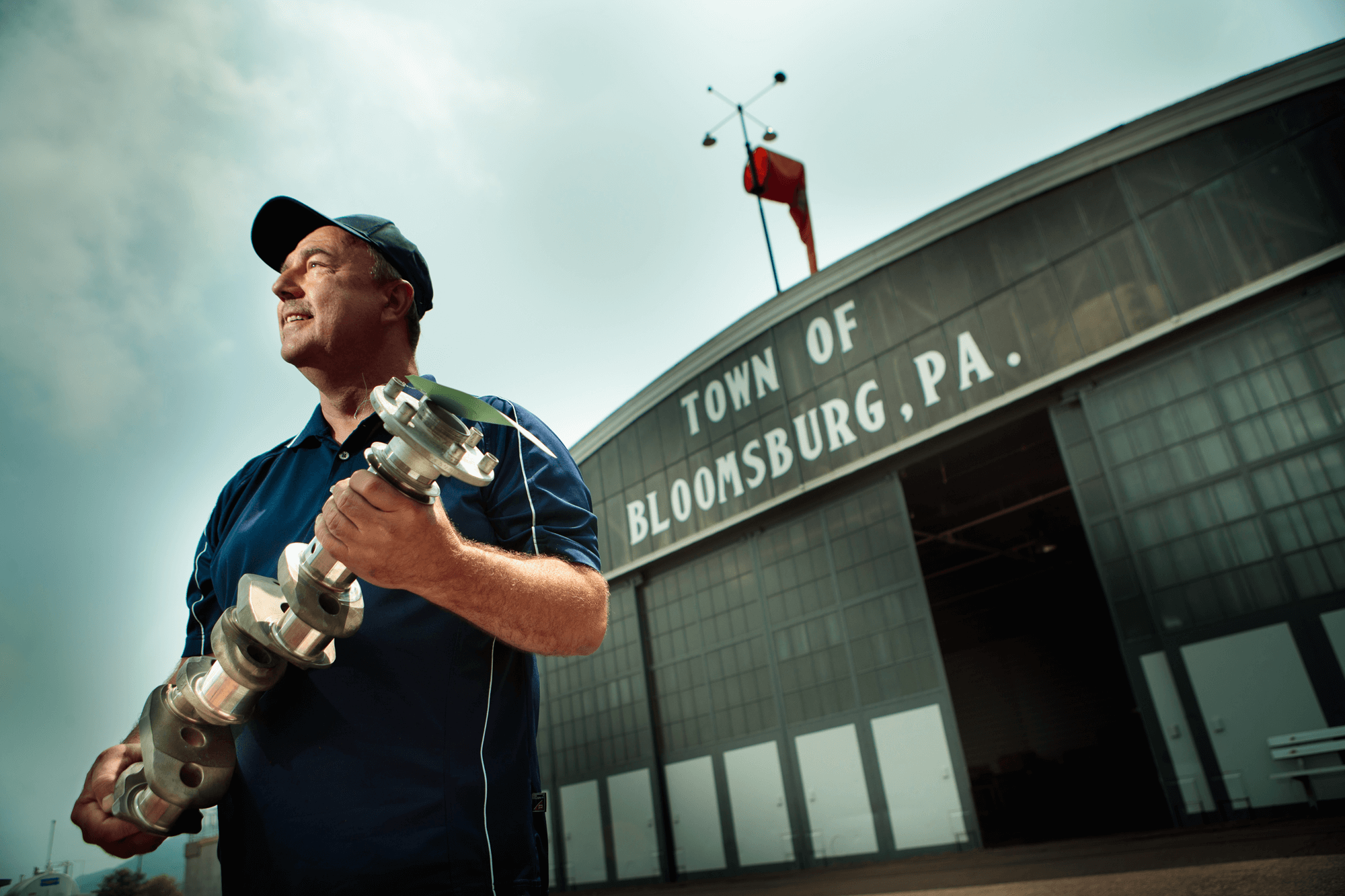 A man holds a machine part in front of an airplane hangar