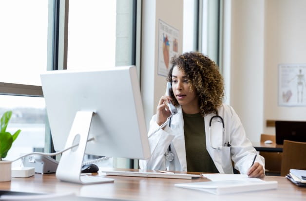 Focused female doctor on phone call, using computer, medical notes. Healthcare professional at work.