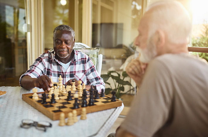 Two older men sit at a table outside playing chess