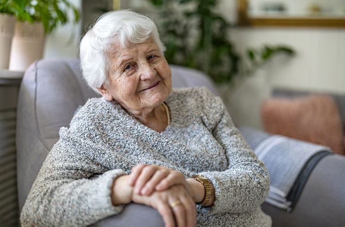 an older woman sits in a chair in her home smiling at the camera
