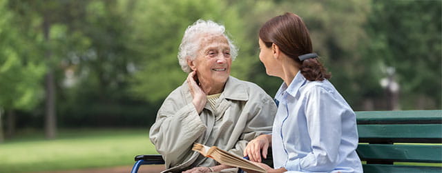 an older woman sits on a park bench smiling at a younger woman who is talking to her