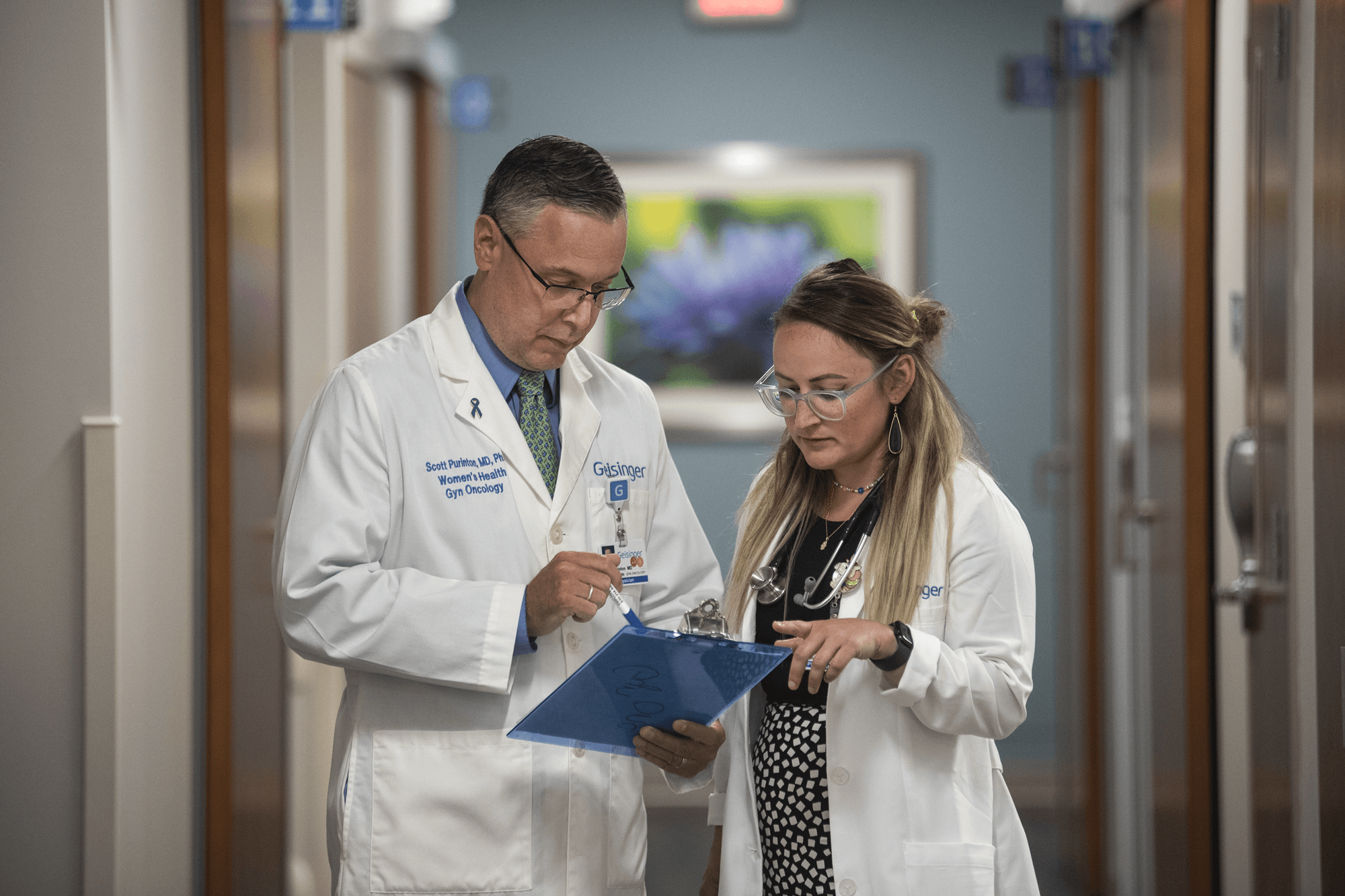 Two doctors stand in a hallway discussing results on a clipboard