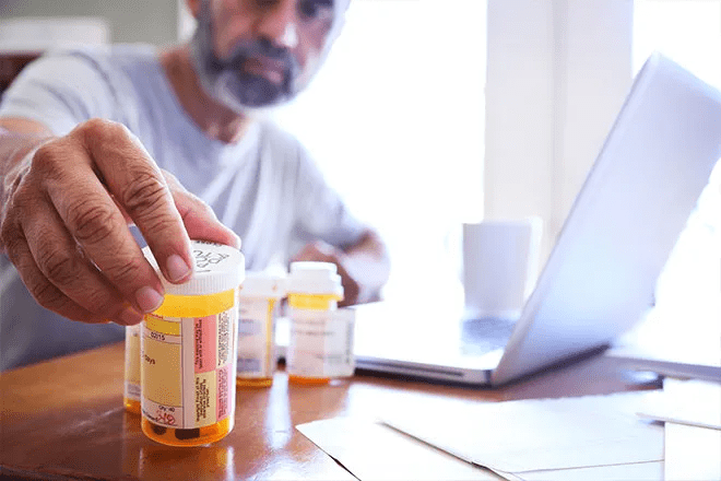 A senior sitting at a desk with a laptop looks at his prescription bottle