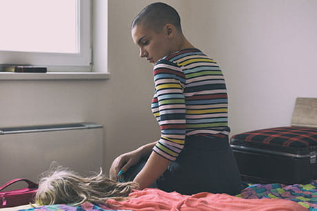 young woman with shaved head sitting pensively with her wig in her hands