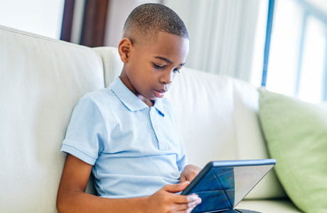 a young boy sits on the couch looking at a tablet