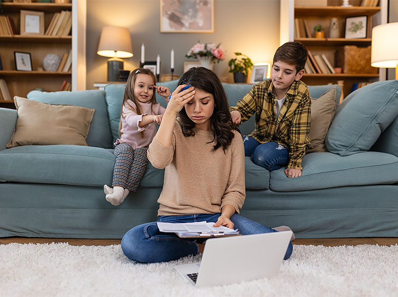 Person reviewing paperwork on a laptop while caring for children at home, illustrating the challenges of balancing family responsibilities and daily tasks.
