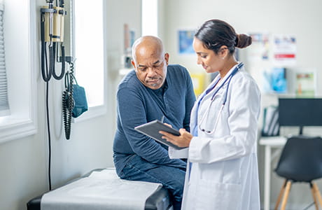 A senior gentleman sits up on an exam table. A female doctor is holding out a tablet as they review test results together.