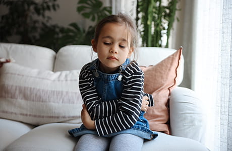 A young child sits on a couch with her arms crossed