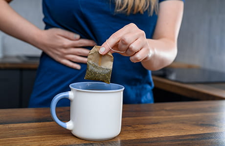 a woman pulling a teabag out of a cup while holding her stomach