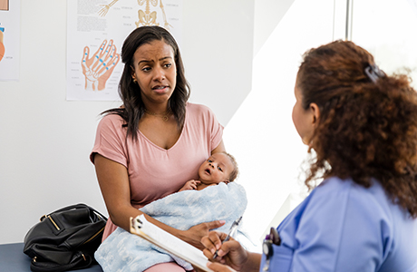 A mother with a tiny baby sits in a doctors office talking about postpartum depression with a female doctor