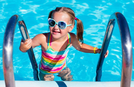 A young girl in a rainbow-colored bathing suit, climbs out of a swimming pool.