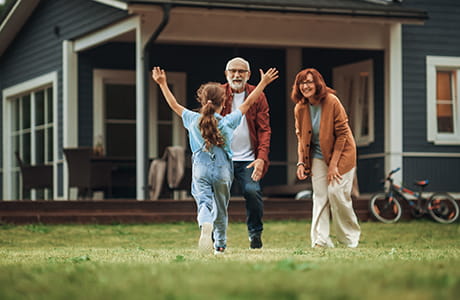 Child runs across a lawn toward two older adults standing outside a house.