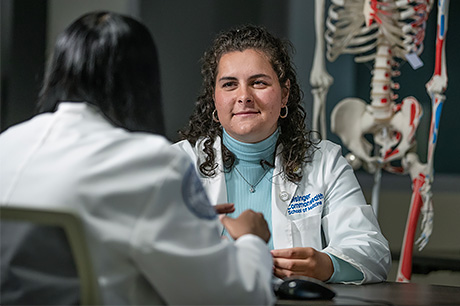 Two individuals in white coats by a desk with a skeleton model in the background.