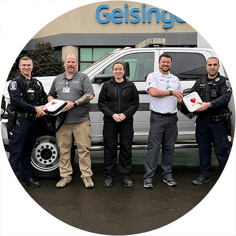 First responders and healthcare team members standing outside a Geisinger facility with automated external defibrillators and an emergency medical services vehicle.