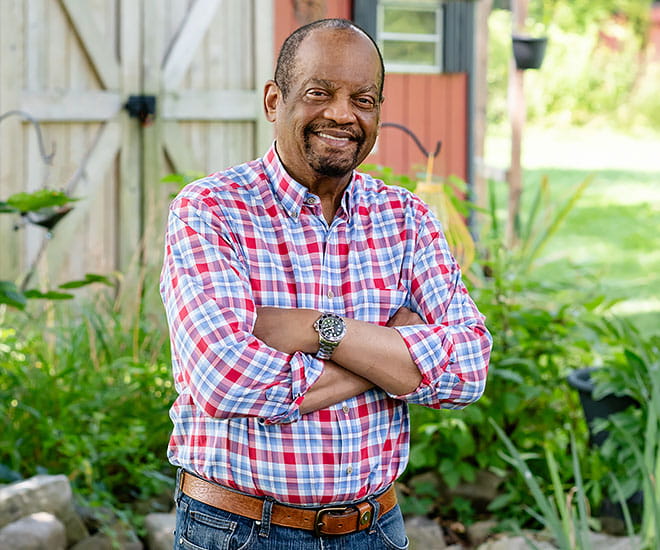 Person standing outdoors in a garden setting with arms crossed among plants and greenery.